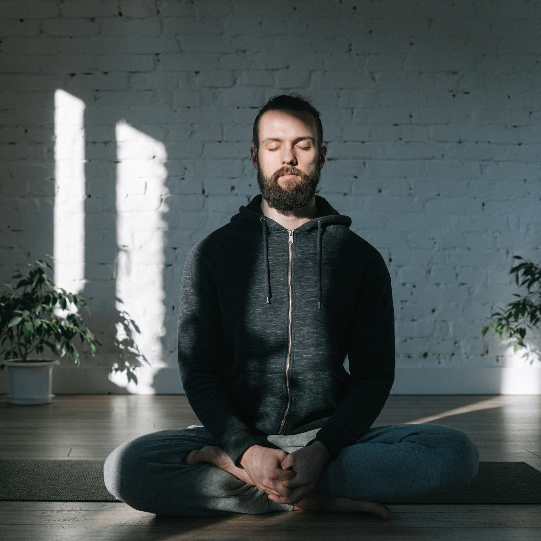 A man with a beard sits cross-legged on a yoga mat in a bright room with a white brick wall, wearing a dark gray zip-up hoodie and gray sweatpants, with sunlight streaming in from a window.