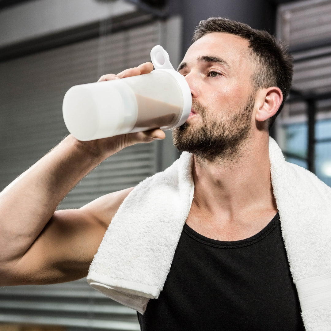 A man in a gym setting drinking from a white plastic shaker bottle with a white towel draped over his shoulders.