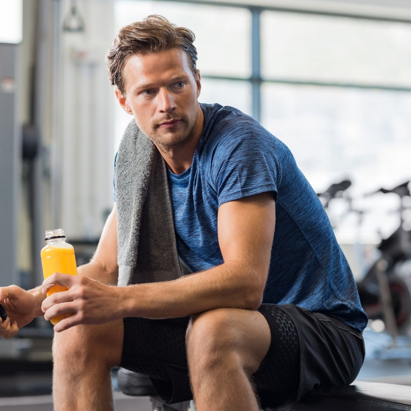 A man in a gym setting wearing a blue athletic t-shirt and black shorts, sitting on a bench with a gray towel over his shoulder and holding an orange drink.