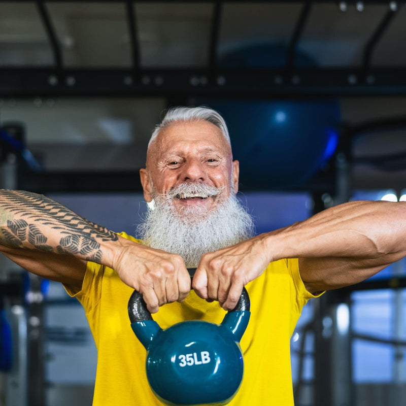 A smiling older man with a white beard and tattooed arm lifts a blue 35LB kettlebell in a gym setting, wearing a bright yellow t-shirt.