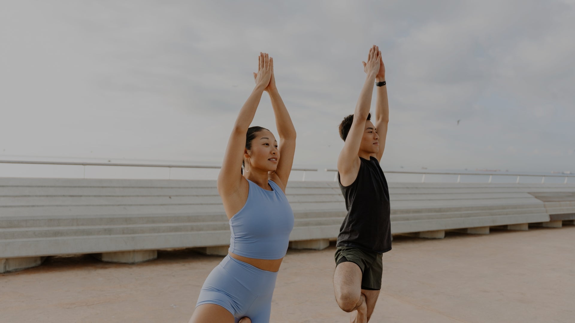 Two people in athletic wear performing a yoga pose outdoors on a cloudy day, with one person in a light blue activewear set and the other in a black sleeveless top and dark shorts.