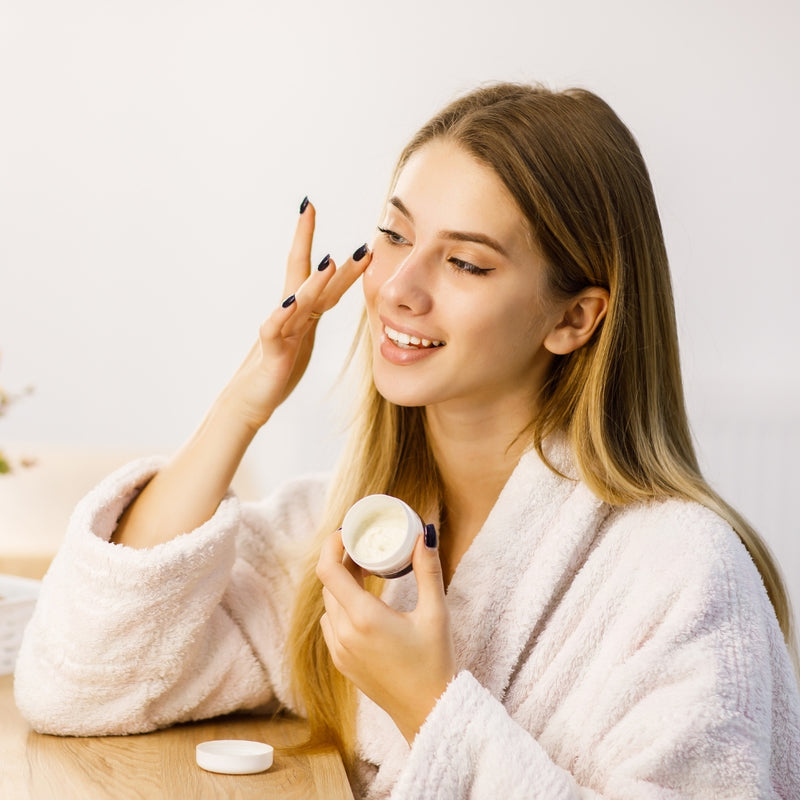 Woman in a white robe applying cream to her face with a neutral background