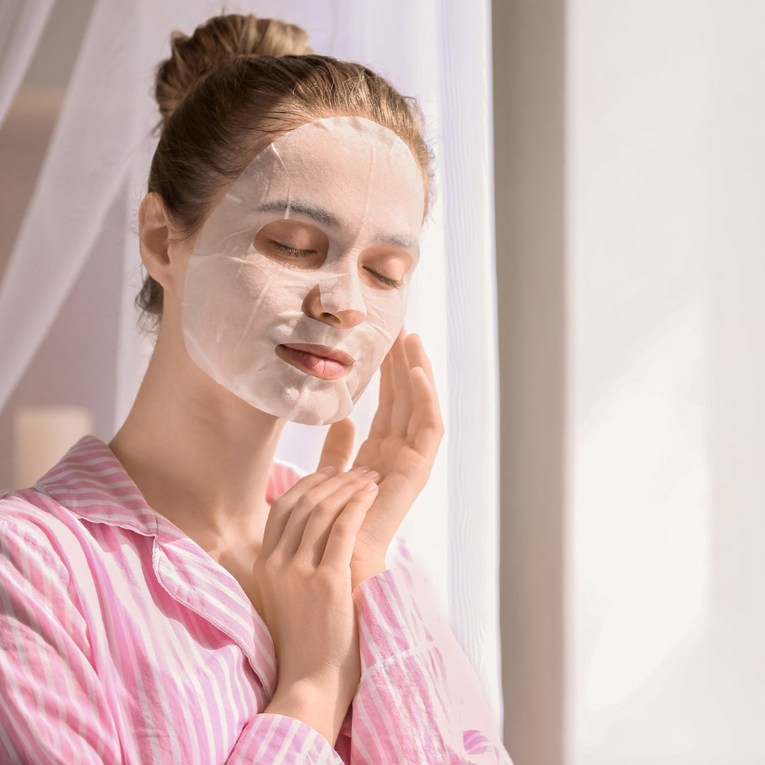 A woman with her hair in a bun applies a translucent sheet mask to her face while wearing pink and white striped pajamas, set against a soft, bright background with sheer curtains.