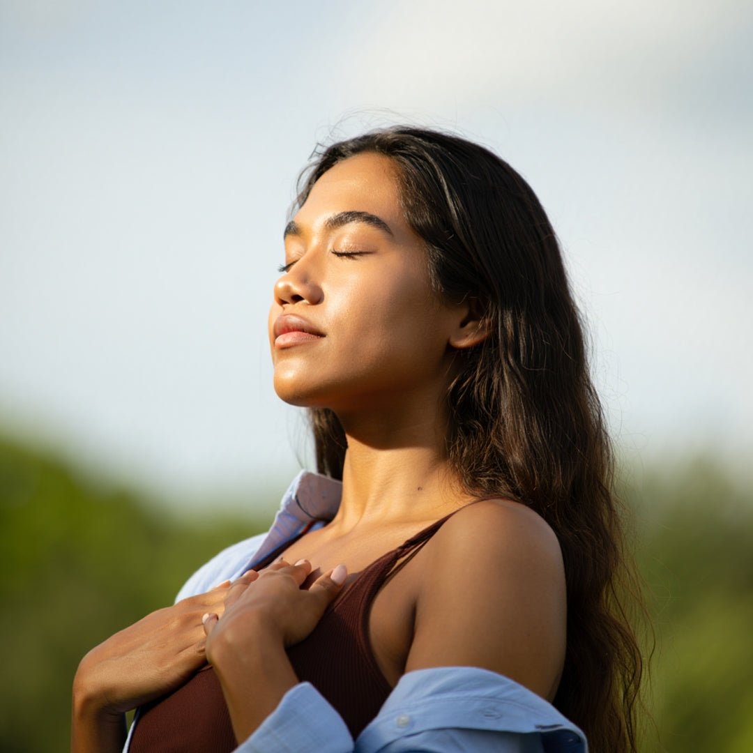 A woman with her eyes closed and hands over her chest, wearing a brown ribbed tank top and a light blue button-up shirt, set against a soft-focus outdoor background.