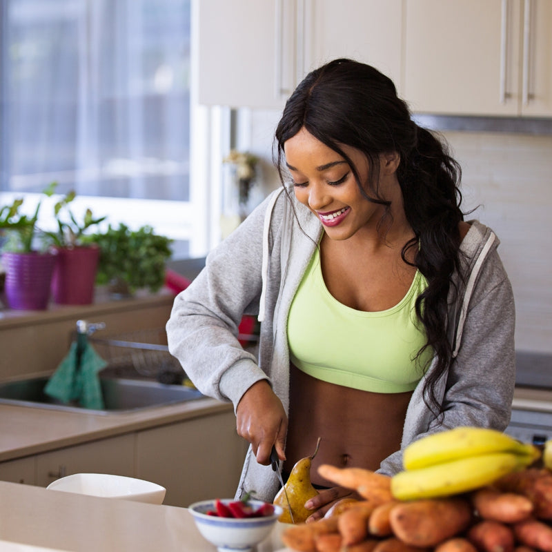 A woman in a kitchen preparing food, wearing a light green sports bra and a gray zip-up hoodie. Bananas and other fruits are visible on the counter in the foreground.